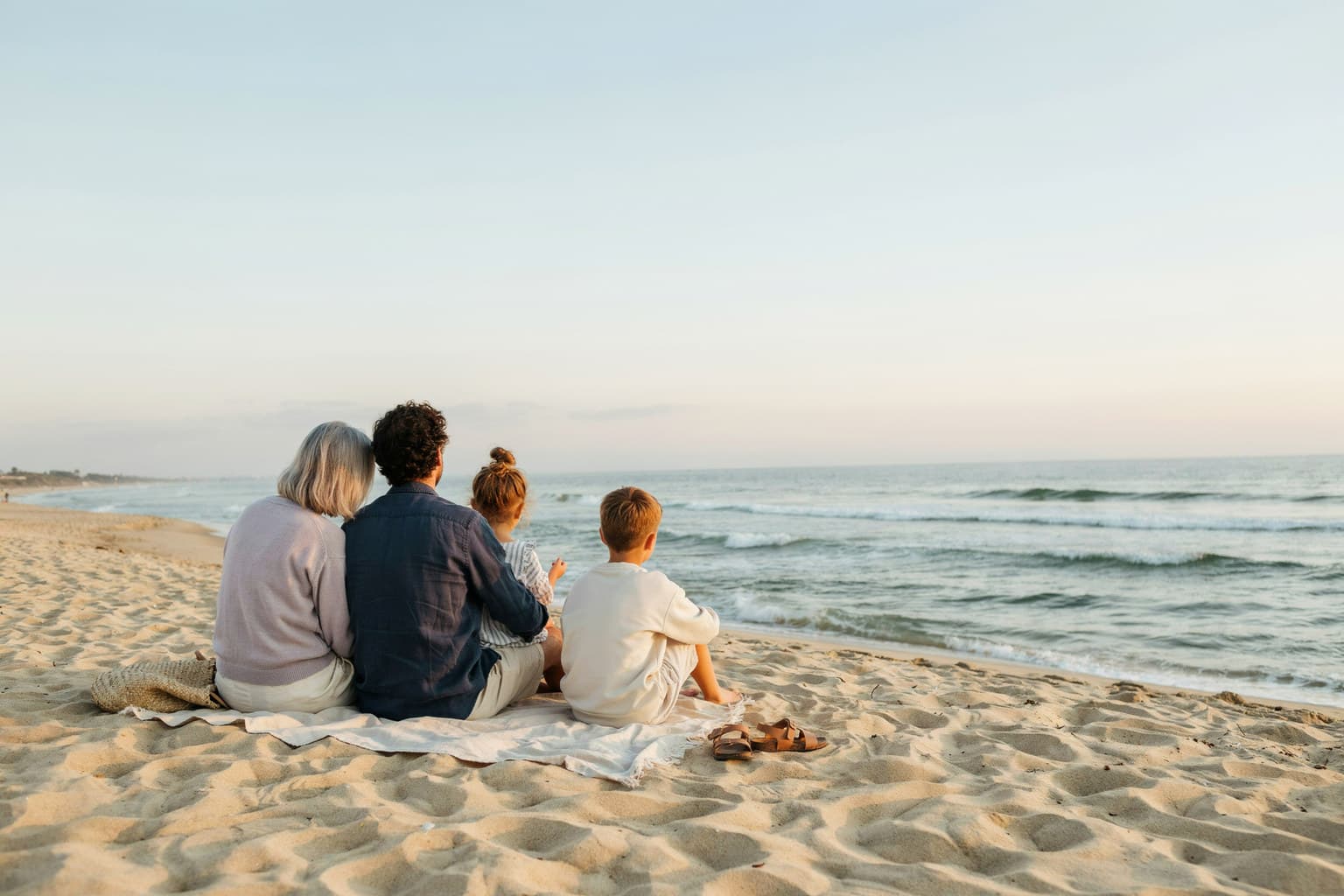 A family sitting together on a blanket on the sand at dusk, looking out at the ocean.